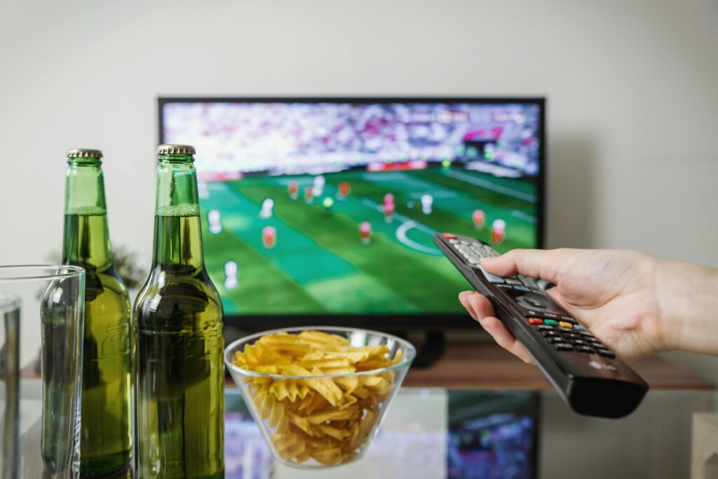 A person holding a remote while watching soccer on TV with beer and snacks on the table.